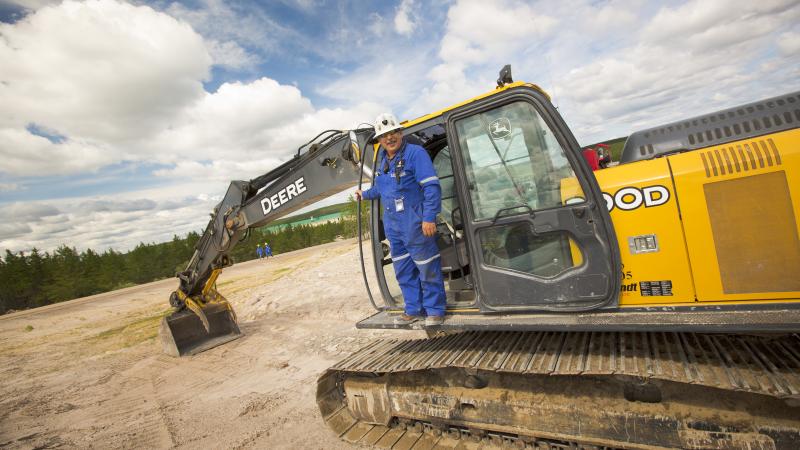 Man in hardhat, sunglasses, and blue coveralls standing on John Deere